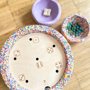 Può includere: Un gioco in legno con illustrazioni di dadi e un bordo colorato, insieme a due ciotole. Una ciotola è viola e contiene dadi, mentre l'altra è decorata con perline colorate. Il tabellone ha una finitura in legno chiaro.