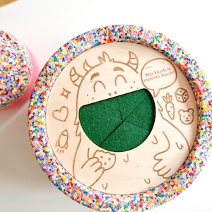 Op de afbeelding: Een houten speelgoed met een monsterillustratie en de tekst "Was kitzelt in meinem Bauch?". Het speelgoed heeft een groene vilten inzet en een kleurrijke, gespikkelde rand. Andere illustraties zijn een hart, een raket, een wortel, een dobbelsteen en een beer.
