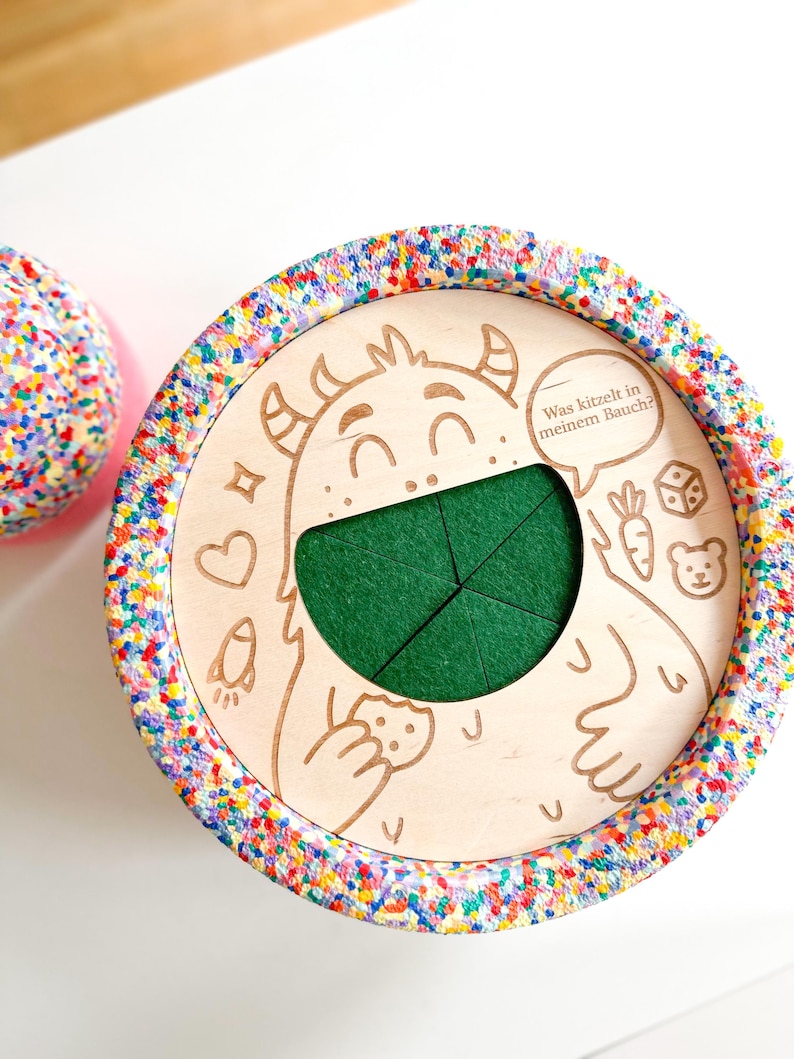 K&ouml;nnte beinhalten: Ein Holzspielzeug mit einer Monsterillustration und dem Text "Was kitzelt in meinem Bauch?". Das Spielzeug hat einen gr&uuml;nen Filzeinsatz und einen bunten, gesprenkelten Rand. Weitere Illustrationen sind ein Herz, eine Rakete, eine Karotte, ein W&uuml;rfel und ein B&auml;r.