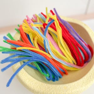Op de afbeelding: Een verzameling kleurrijke pijpenragers in een lichtgele kom. De pijpenragers zijn in verschillende kleuren, waaronder rood, geel, blauw, groen, oranje, roze en paars. De kom heeft een textuur.