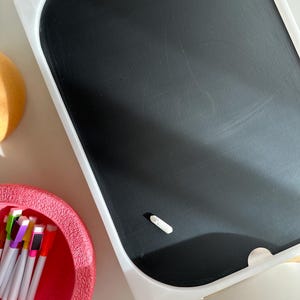Op de afbeelding: Een wit geframed schoolbord met een zwart oppervlak en een klein stukje witte krijt in de rechterbenedenhoek. Het schoolbord staat op een wit oppervlak.