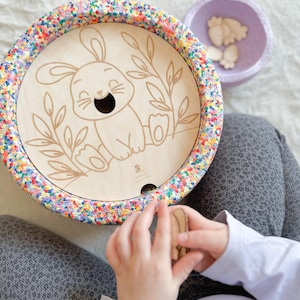 Può includere: Un giocattolo di legno con un bordo colorato e un'illustrazione di un coniglietto sorridente. Il giocattolo ha un foro al centro ed è circondato da motivi floreali. Una piccola ciotola viola con zampine di coniglio in legno è vicina.