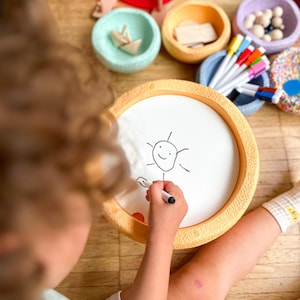 Può includere: Un bambino disegna un sole su una lavagna bianca con un pennarello nero. La lavagna ha una cornice gialla strutturata. Varie ciotole colorate con giocattoli di legno e pennarelli sono disposte intorno alla lavagna su un pavimento di legno.