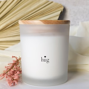 May include: White frosted glass candle jar with a wooden lid. The candle has a black heart and the word "hug" printed on the front.