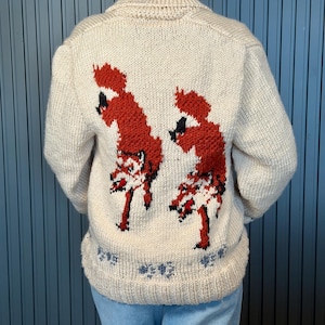 May include: Cream-colored knit sweater with a high collar and long sleeves. The back features two red fox designs with black accents. The bottom hem has a blue floral pattern. A cozy, warm sweater for fall and winter.