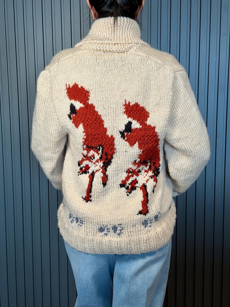 May include: Cream-colored knit sweater with a high collar and long sleeves. The back features two red fox designs with black accents. The bottom hem has a blue floral pattern. A cozy, warm sweater for fall and winter.