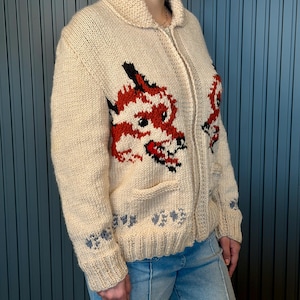 May include: Cream-colored, knitted zip-up sweater featuring a red and black dragon design on the front. The sweater has a shawl collar, two front pockets, and blue floral accents on the cuffs and hem. The model is wearing light blue jeans.