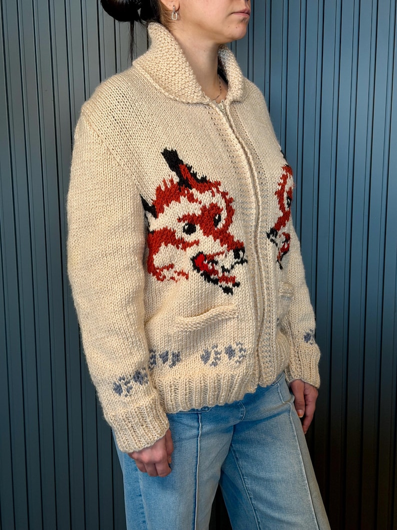 May include: Cream-colored, knitted zip-up sweater featuring a red and black dragon design on the front. The sweater has a shawl collar, two front pockets, and blue floral accents on the cuffs and hem. The model is wearing light blue jeans.