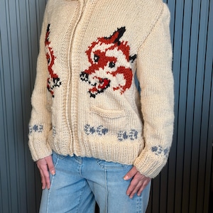 May include: Cream-colored, zip-up knit sweater with a dragon design in red, black, and white. The sweater features a collared neckline, two pockets, and blue floral accents on the sleeves and hem. The dragon is on the front of the sweater.
