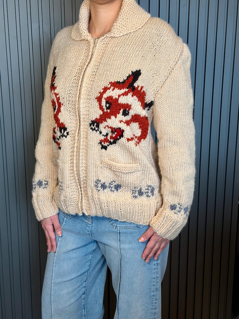 May include: Cream-colored, zip-up knit sweater with a dragon design in red, black, and white. The sweater features a collared neckline, two pockets, and blue floral accents on the sleeves and hem. The dragon is on the front of the sweater.