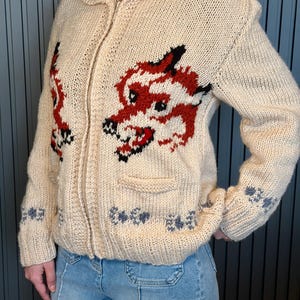 May include: Cream-colored, zip-up knit sweater with a red and black dragon design on the front. The sweater has a collared neckline, a small pocket, and decorative blue and gray floral patterns on the sleeves and bottom hem.