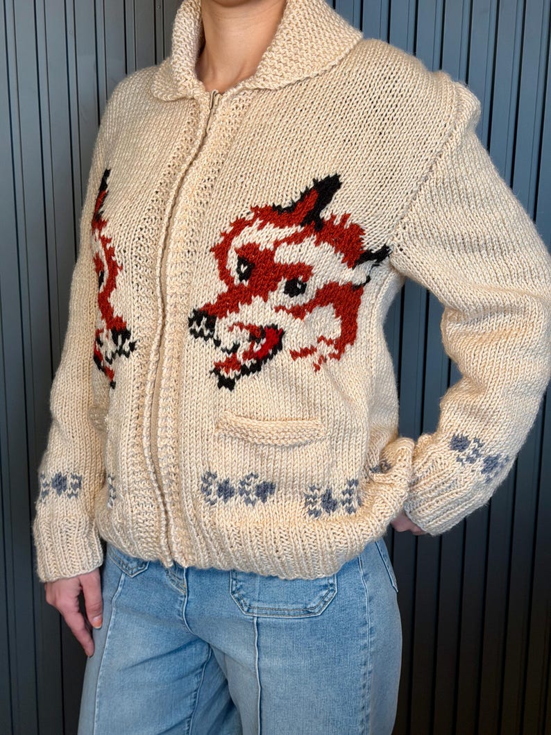May include: Cream-colored, zip-up knit sweater with a red and black dragon design on the front. The sweater has a collared neckline, a small pocket, and decorative blue and gray floral patterns on the sleeves and bottom hem.