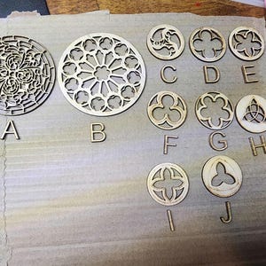 May include: A collection of laser-cut wooden decorative elements, including circular and geometric shapes. The designs feature intricate patterns and shapes, labeled with letters A through J. The pieces are arranged on a light brown surface.