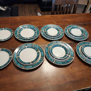 May include: A collection of eight ceramic plates and bowls featuring a white center and a wide, teal-colored border with a marbled pattern and thin black geometric lines. The set includes various sizes of round dinnerware arranged in two rows.