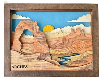 Puede incluir: Una obra de arte de madera en capas que representa un paisaje desértico con un gran arco, montañas y un río. El cielo es azul con nubes blancas y un sol amarillo. La palabra "ARCHES" está impresa en la parte inferior.
