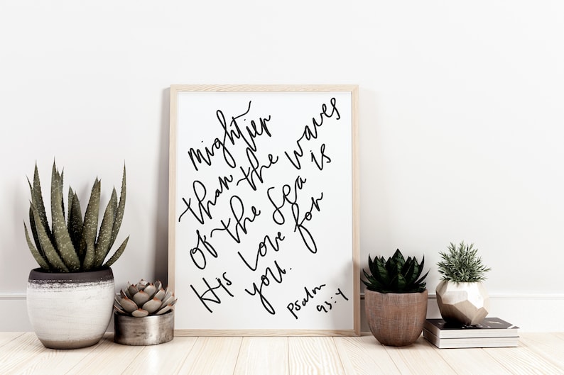 May include: A framed print with a white background and black calligraphy text that reads "mightier than the waves of the sea is His love for you. Psalm 93:4". The print is sitting on a wooden floor with two potted plants and a stack of books.
