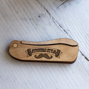 Op de afbeelding: Een houten kam met een gebogen handvat en de tekst "Hipsters Style" met een snor-afbeelding.