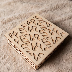 May include: A square wooden box with a geometric pattern cut out of the top. The box is made of light wood and has a natural finish.