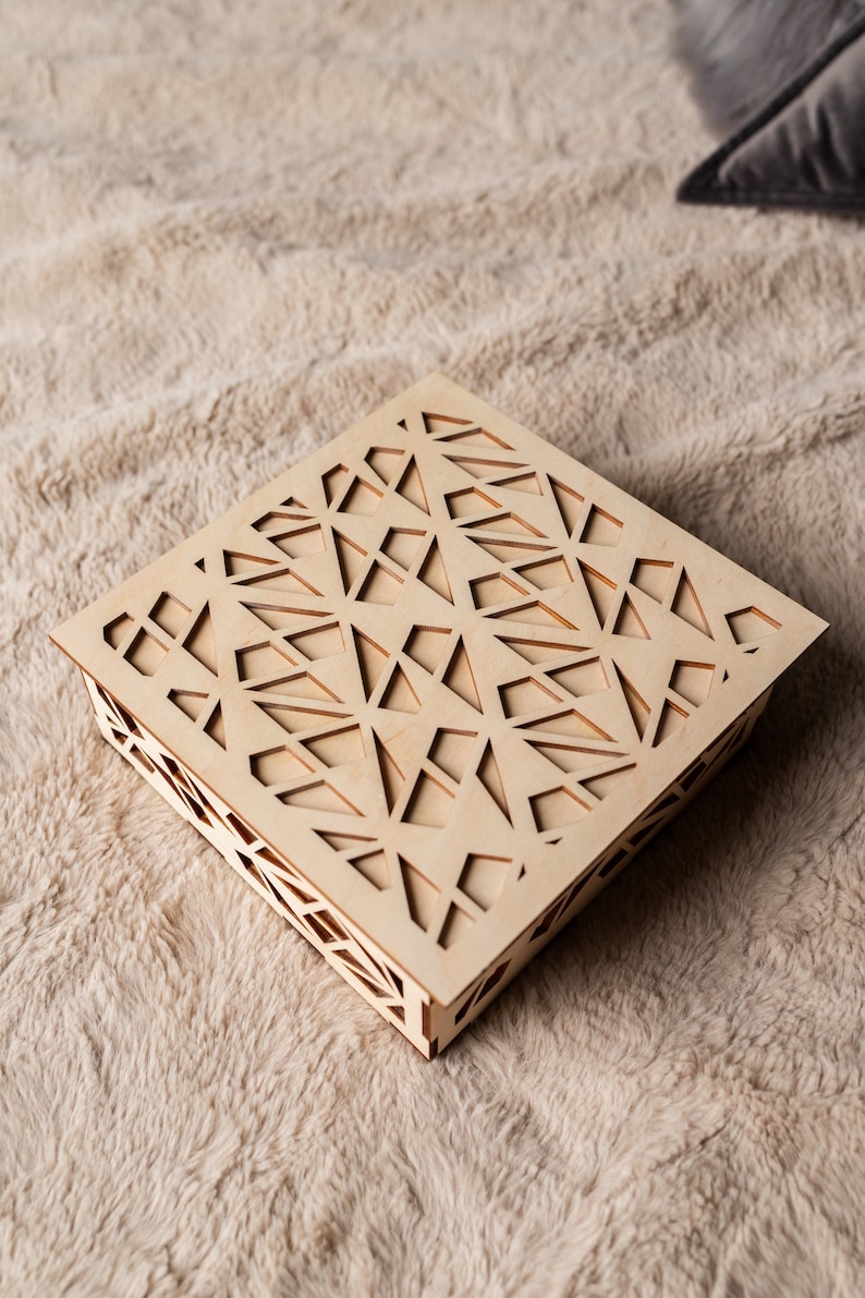 May include: A square wooden box with a geometric pattern cut out of the top. The box is made of light wood and has a natural finish.