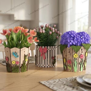 Può includere: Tre vasi di fiori decorativi in legno con fiori colorati. I vasi hanno un design a forma di recinzione con fiori, farfalle ed erba ritagliati. I fiori includono tulipani, fiori rosa e ortensie viola.