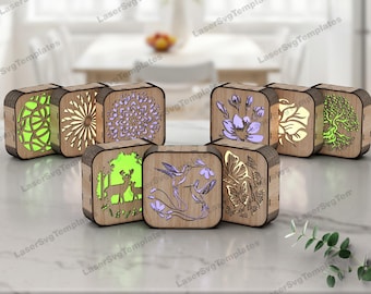 May include: A collection of wooden, laser-cut boxes with various nature-themed designs. Each box features intricate cutouts of a tree, flowers, hummingbirds, deer, and other patterns. The boxes are illuminated with different colored lights, creating a warm glow.