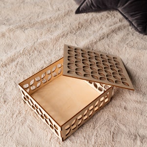 May include: An open wooden box with a geometric pattern cut out of the sides and lid. The box is empty and sitting on a light brown fuzzy surface.