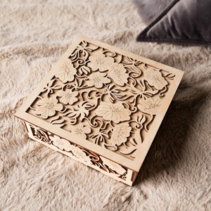 May include: A wooden box with a delicate floral design carved into the lid. The box is light brown and has a square shape.