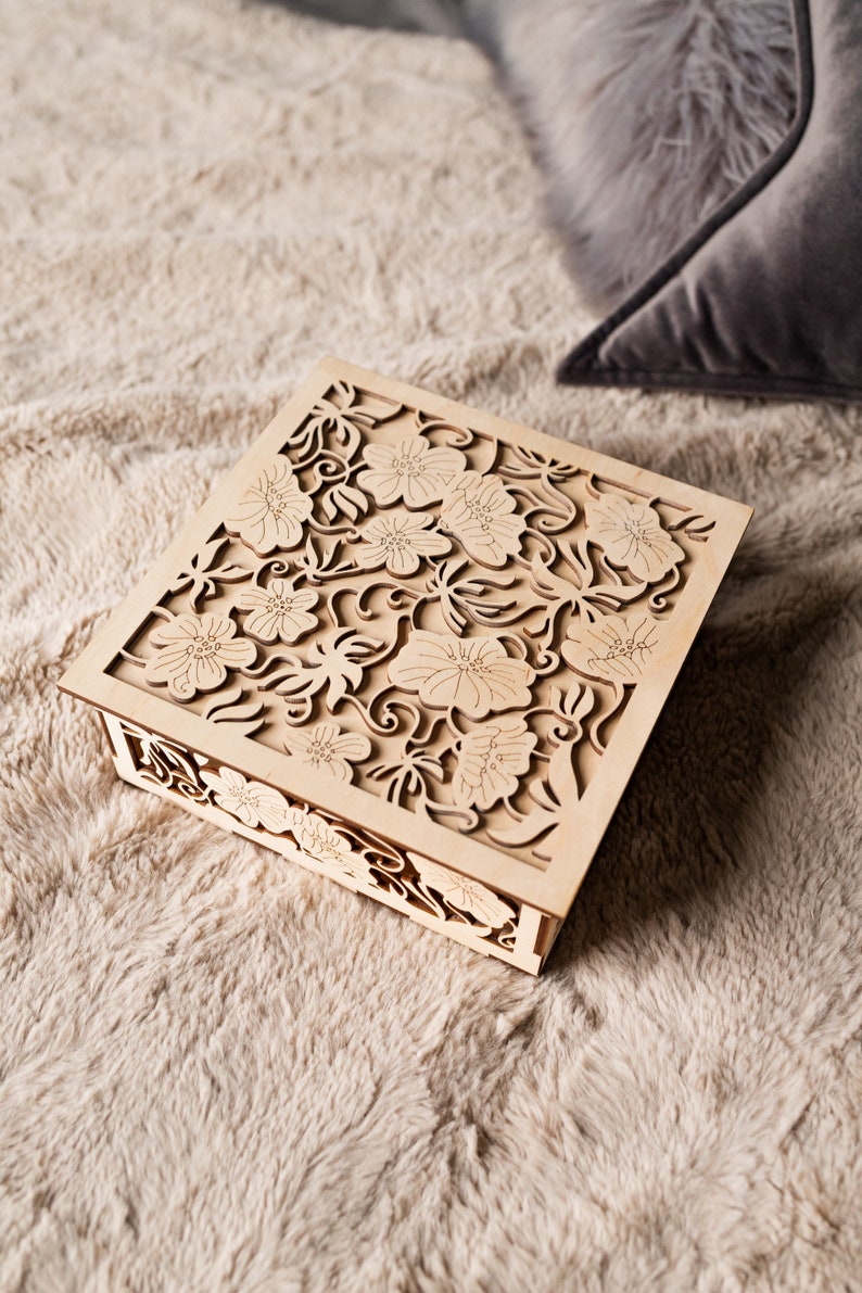 May include: A wooden box with a delicate floral design carved into the lid. The box is light brown and has a square shape.