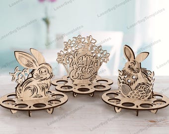May include: Wooden Easter egg holder with three different designs. One design features a bunny with flowers, another features a bunny with eggs, and the last design features a basket with flowers and the text "Happy Easter".