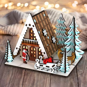 May include: A wooden Christmas village scene with a gingerbread house, two pine trees, a reindeer, and Santa Claus. The house has a brown roof, white trim, and windows with glowing lights. The trees are decorated with blue and white snowflakes. The reindeer is brown with antlers. Santa Claus is wearing a red suit and a white beard. The scene is set on a white base.
