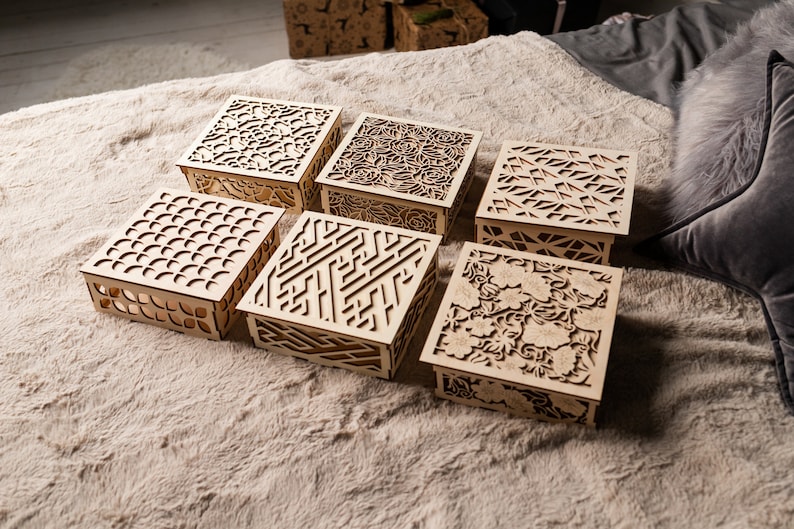 May include: Six wooden boxes with intricate laser cut designs. The boxes are arranged in a row on a light brown fuzzy surface. The designs include geometric patterns, floral motifs, and abstract shapes.