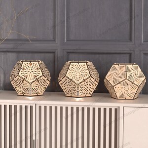May include: Three wooden geometric lamps with intricate cut-out designs. The lamps are illuminated from within, casting a warm glow. The lamps are arranged on a wooden shelf with a vertical slatted design.