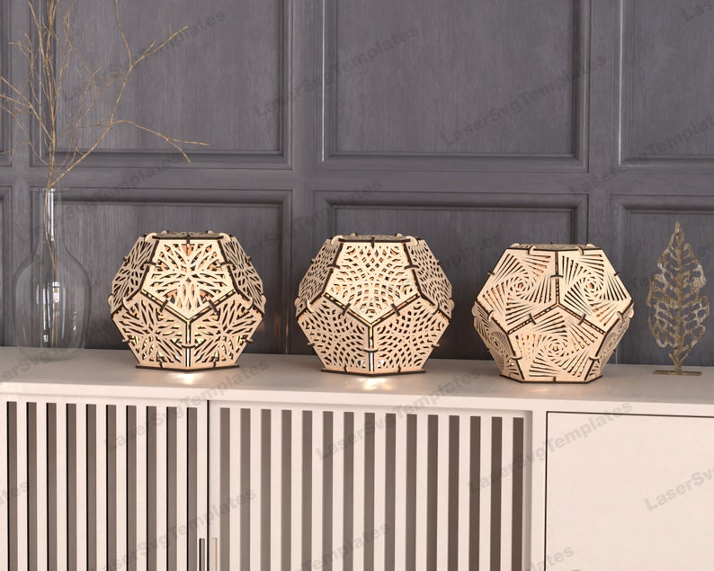 May include: Three wooden geometric lamps with intricate cut-out designs. The lamps are illuminated from within, casting a warm glow. The lamps are arranged on a wooden shelf with a vertical slatted design.