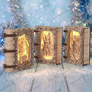 May include: Three rectangular wooden book-shaped light boxes with intricate laser-cut designs. Each box features a warm internal glow illuminating scenes of a nativity, a winter village, and a forest landscape with detailed filigree borders.