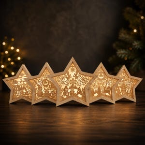 May include: A set of five light-colored wooden star-shaped ornaments, each featuring intricate laser-cut silhouettes of winter scenes including snowmen, reindeer, pine trees, and snowflakes, illuminated from within by a warm glowing light.