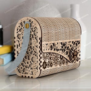 Peut inclure: Un sac à main en bois avec des découpes florales complexes et une bandoulière grise claire. Le sac a un dessus incurvé et une fermeture à rabat avec un motif floral décoratif. Le sac est en bois clair.