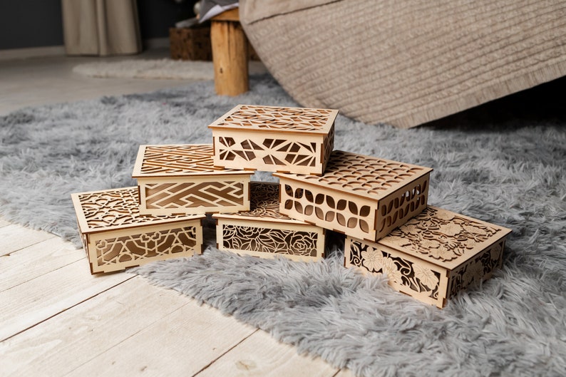 May include: Six wooden boxes with intricate laser cut designs. The boxes are stacked on top of each other on a gray fluffy rug.