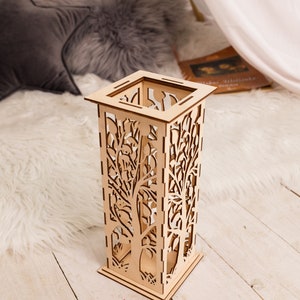 Pode incluir: Um abajur decorativo de madeira com um design de &aacute;rvores recortadas. O abajur &eacute; de cor castanho claro e tem uma forma quadrada.