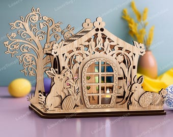 May include: A wooden Easter house with a cutout design, featuring a tree, bunnies, and eggs. The house has a window with a candle inside, creating a warm glow.