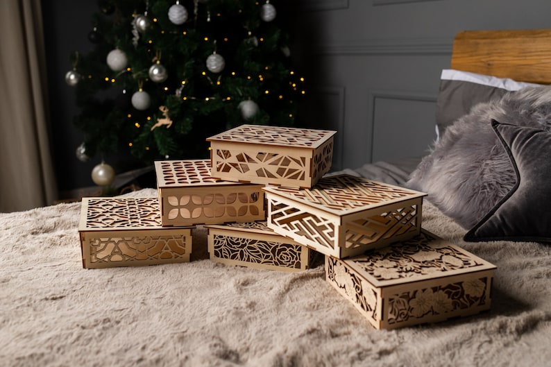 May include: Six wooden boxes with intricate laser-cut designs stacked on a beige surface. The boxes are made of light brown wood and feature various geometric and floral patterns.