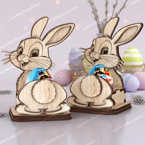 Puede incluir: Dos soportes de madera con forma de conejito de Pascua, cada uno con un diseño recortado, que sostienen un huevo azul y blanco. Los conejitos tienen detalles y están colocados sobre una base marrón. Huevos de Pascua y decoraciones en el fondo.