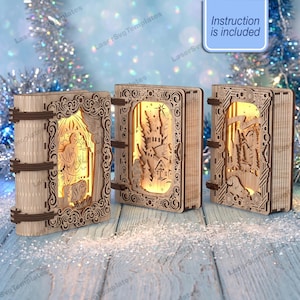 Könnte beinhalten: Drei beleuchtete, buchförmige Holzornamente mit filigranen Laserschnitt-Szenen einer Krippe, eines Winterdorfes und einer Waldlandschaft. Jedes Stück hat einen hellen Holzrahmen mit dunkelbraunen Akzenten und ein blaues Schild mit der Aufschrift Instruction is included.