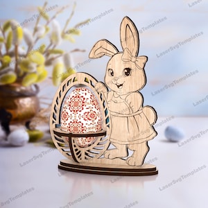 May include: Wooden Easter bunny figurine holding an egg-shaped holder with a decorative egg. The bunny has a bow and dress. The egg has a red and white floral pattern. The figurine is made of light brown wood.