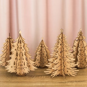 May include: Five wooden Christmas tree decorations in various sizes and designs. The trees are made of laser-cut wood and have intricate details. They are arranged on a wooden surface.