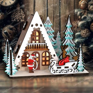 May include: A wooden Christmas village scene with a triangular house, a Santa Claus figure, a sleigh with a red bag, and several pine trees. The house has windows with glowing lights and a white roof with a scalloped edge.