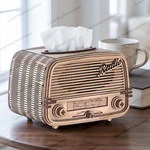 May include: A light-coloured wooden tissue box cover shaped like a vintage radio with a horizontal slatted front, three circular knobs, a printed frequency dial, and the word Radio in script. White tissues protrude from the top opening.