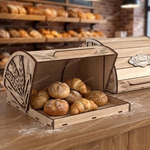 May include: A wooden bread box with a hinged lid, filled with various types of bread, including rolls and croissants. The box has a wheat stalk design on the side and is sitting on a wooden surface.