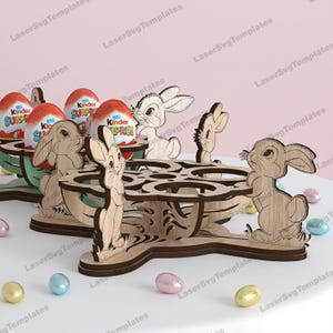 Può includere: Portauova di Pasqua in legno con intagli a forma di coniglietto. I portauova sono progettati per contenere uova di cioccolato. L'immagine mostra diversi portauova, alcuni con uova Kinder Sorpresa. Uova di Pasqua dai colori pastello sono sparse.