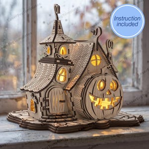 Può includere: Un modello di casa stregata in legno tagliato al laser con una grande facciata a forma di zucca intagliata, un interno luminoso, diverse finestre, una torre alta e un tetto strutturato. La struttura poggia su una base con un'etichetta blu Instruction included.