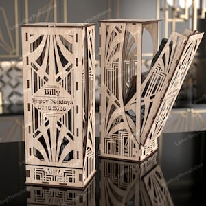 Puede incluir: Dos cajas de vino de madera de estilo Art Déco. Una caja está cerrada, la otra está abierta, revelando una botella en su interior. Las cajas tienen intrincados cortes geométricos y el texto "Billy Happy Holidays 07.10.2026."